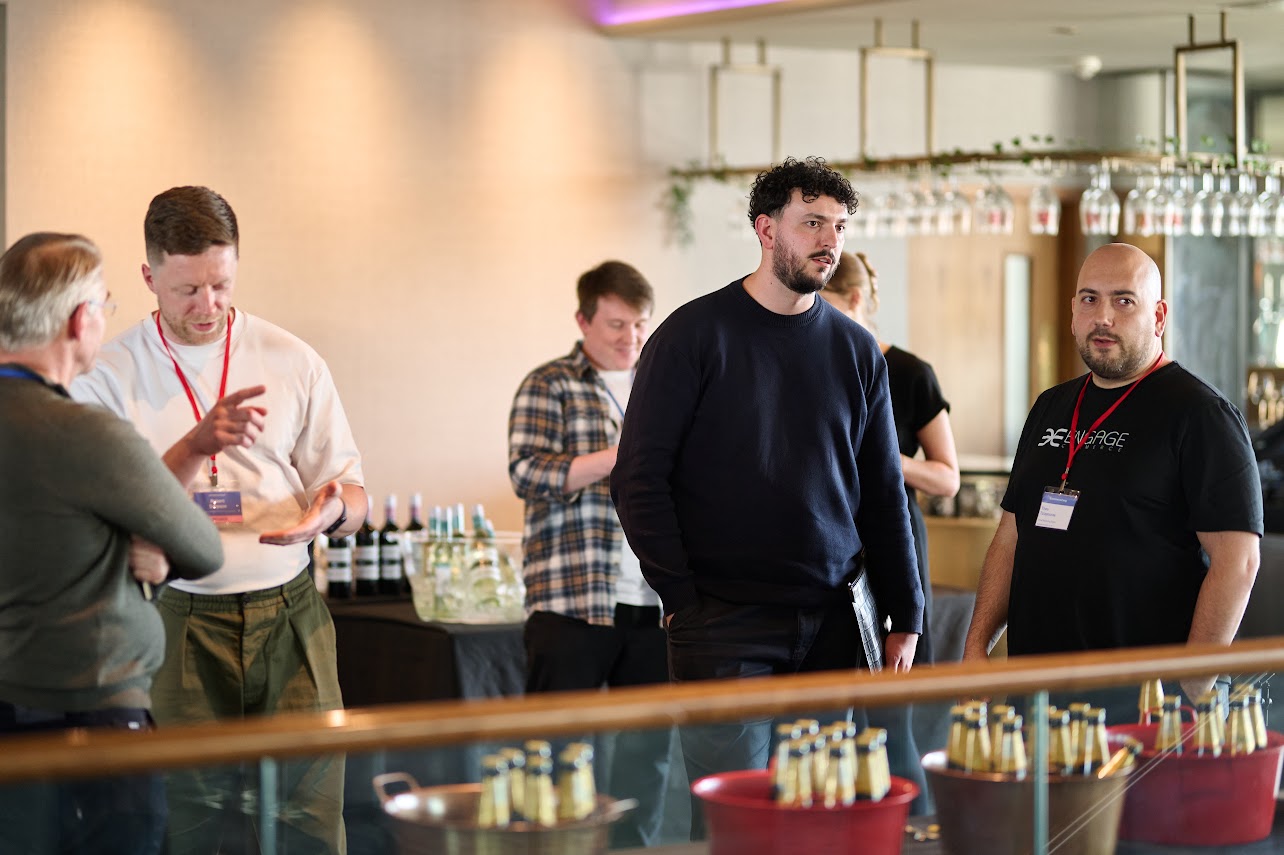 Theo Tziapouras, on the right in a branded Engage Commerce t-shirt, in conversation with delegates beside the drinks table at EcommerceCamp