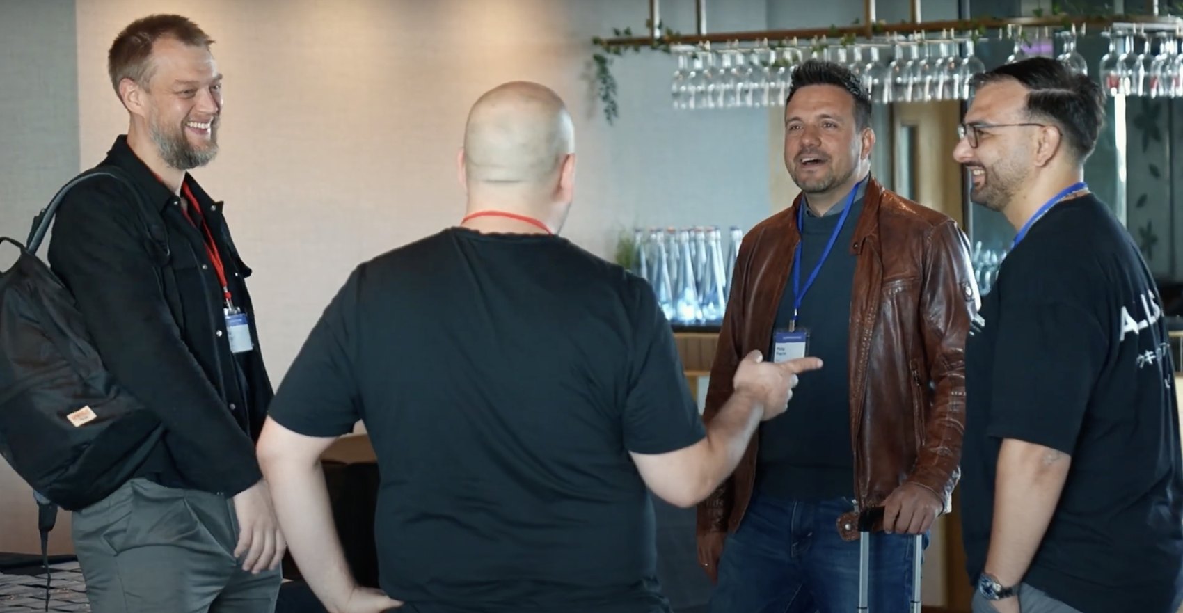 Four delegates laughing in conversation at a drinks reception, lanyards on, the bar and glassware behind them