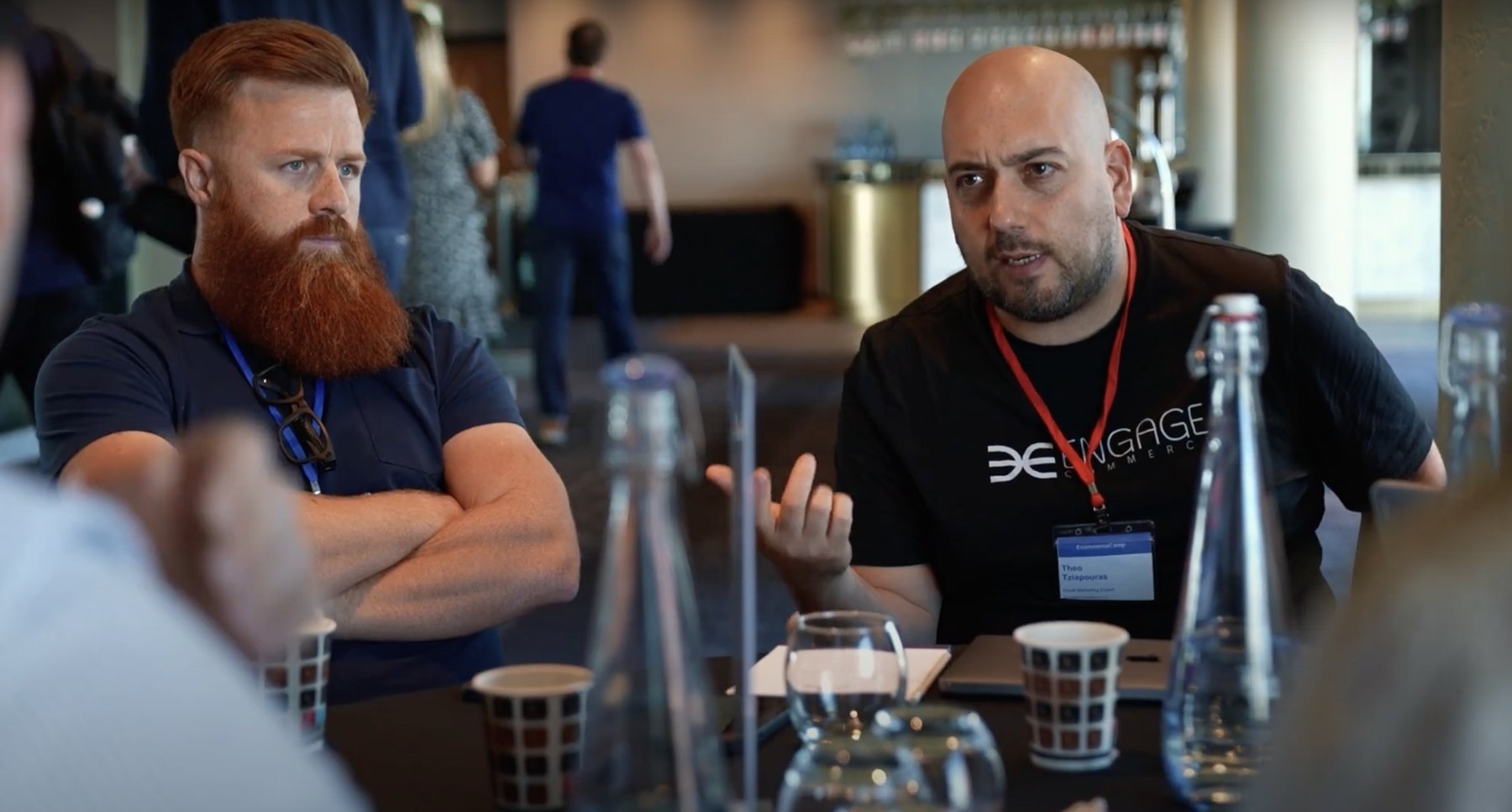 Theo Tziapouras in a black Engage Commerce t-shirt and EcommerceCamp lanyard, mid-sentence across a table from a delegate with a laptop and water bottles on it