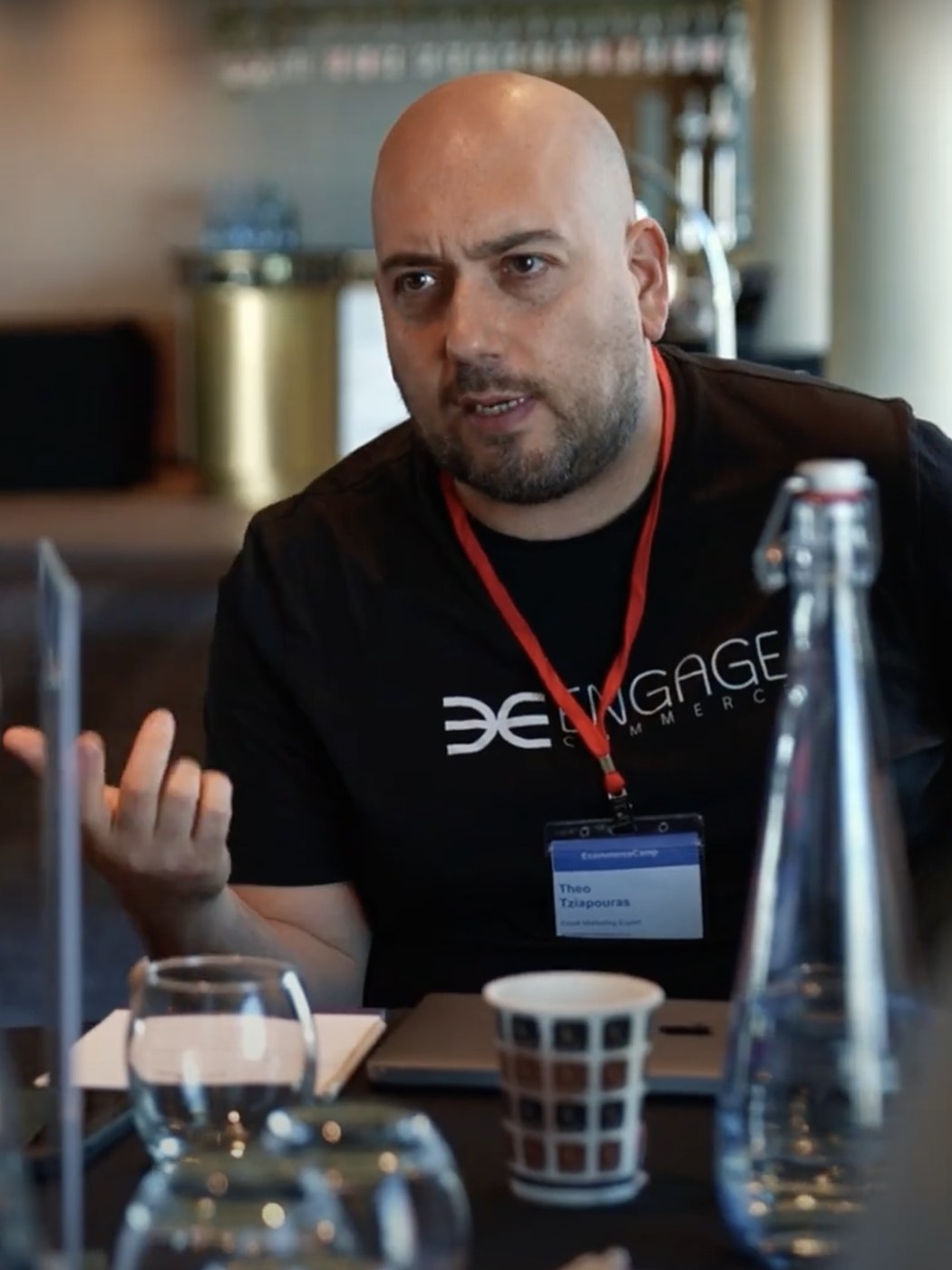 Theo Tziapouras in a black Engage Commerce t-shirt and lanyard, mid sentence across a table at an email roundtable
