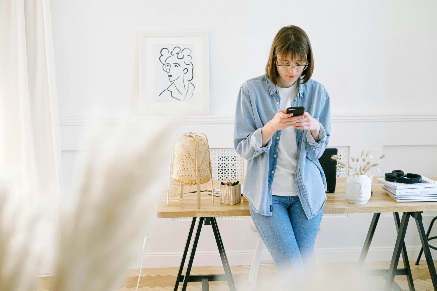 A woman in a denim shirt leaning against a wooden trestle desk in a bright white room, reading something on her phone