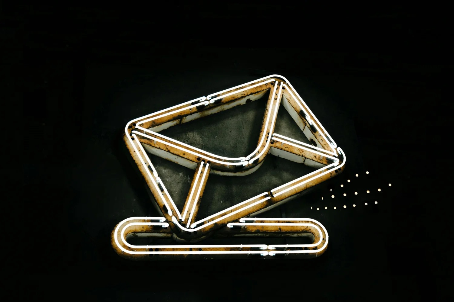 A neon sign in the shape of an envelope glowing white and amber against a black wall, with a trail of small dots leaving one corner