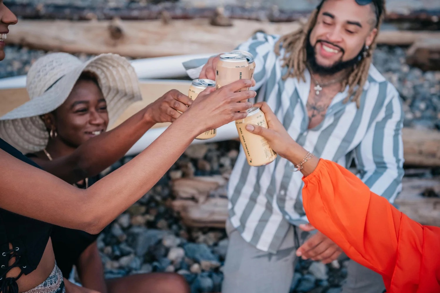 Friends on a rocky beach clinking pale drinks cans together, one woman in a wide sun hat and a man in a striped summer shirt