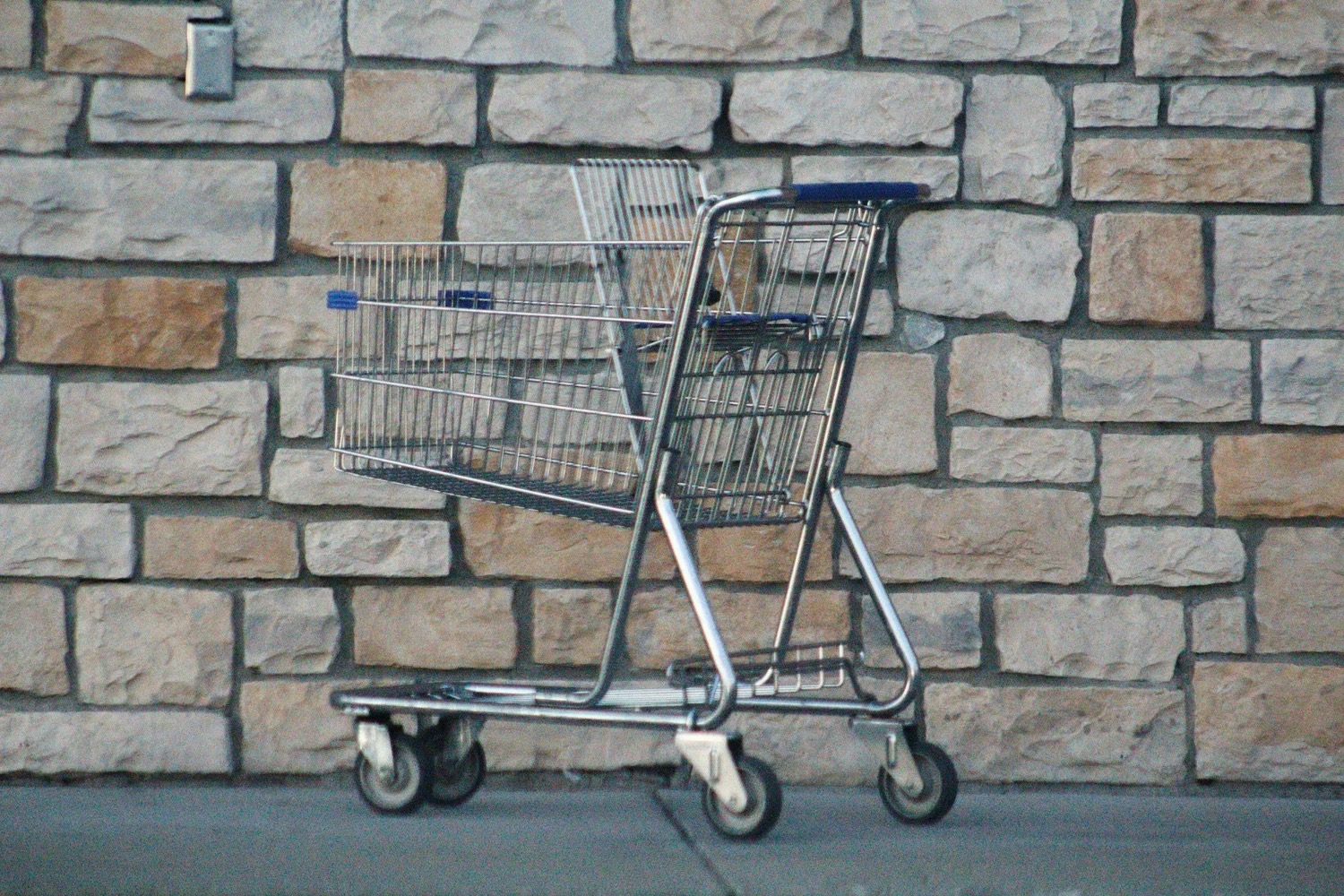 An empty metal shopping trolley with blue handles standing on a pavement in front of a rustic beige stone wall