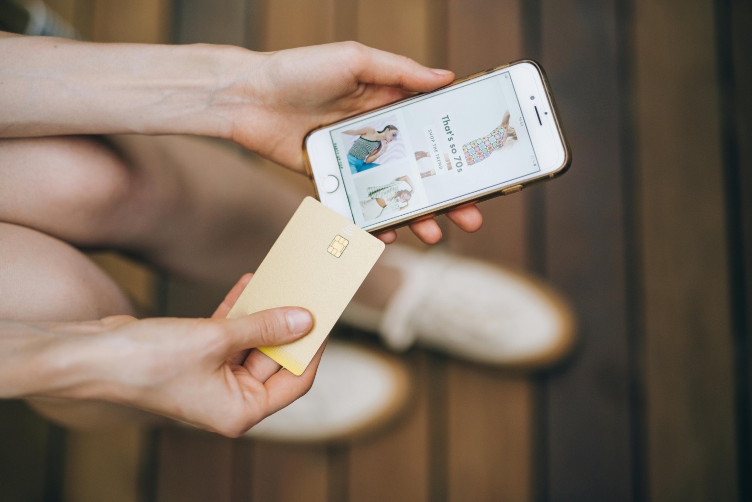 A woman sitting on a wooden bench holding a phone open on a clothing shop app in one hand and a gold bank card in the other