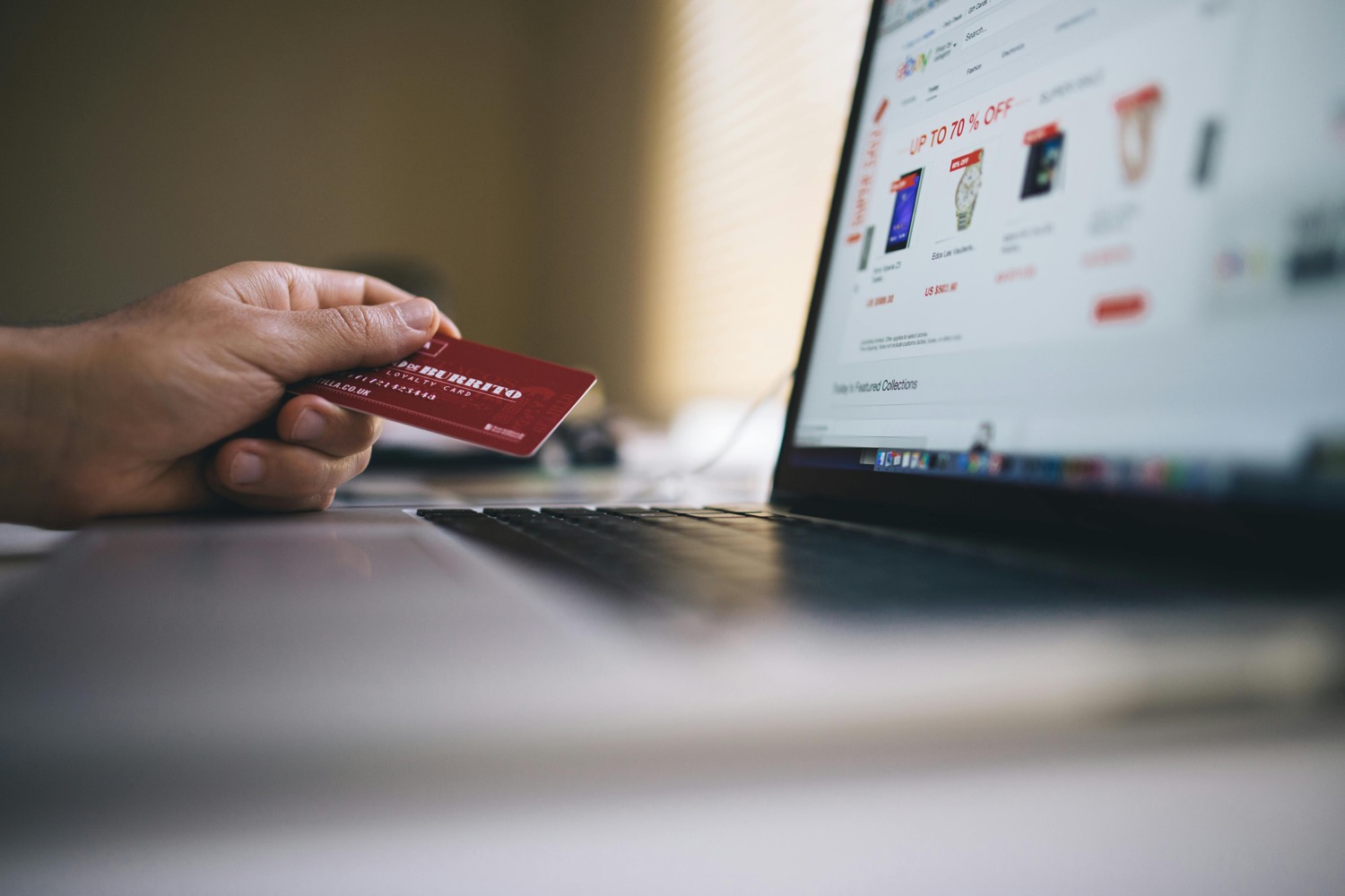 A hand holding a red loyalty card in front of an open laptop showing an online shop's sale listings