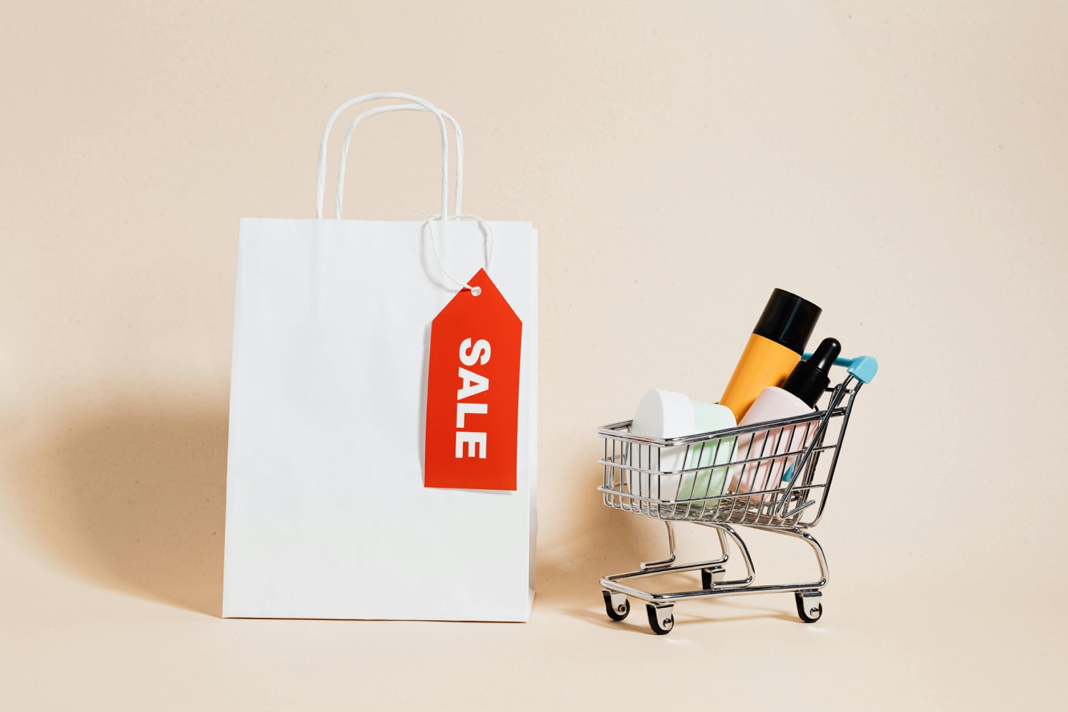 A white paper shopping bag with a red sale tag hanging from it beside a miniature wire trolley holding cosmetics, on a warm cream background