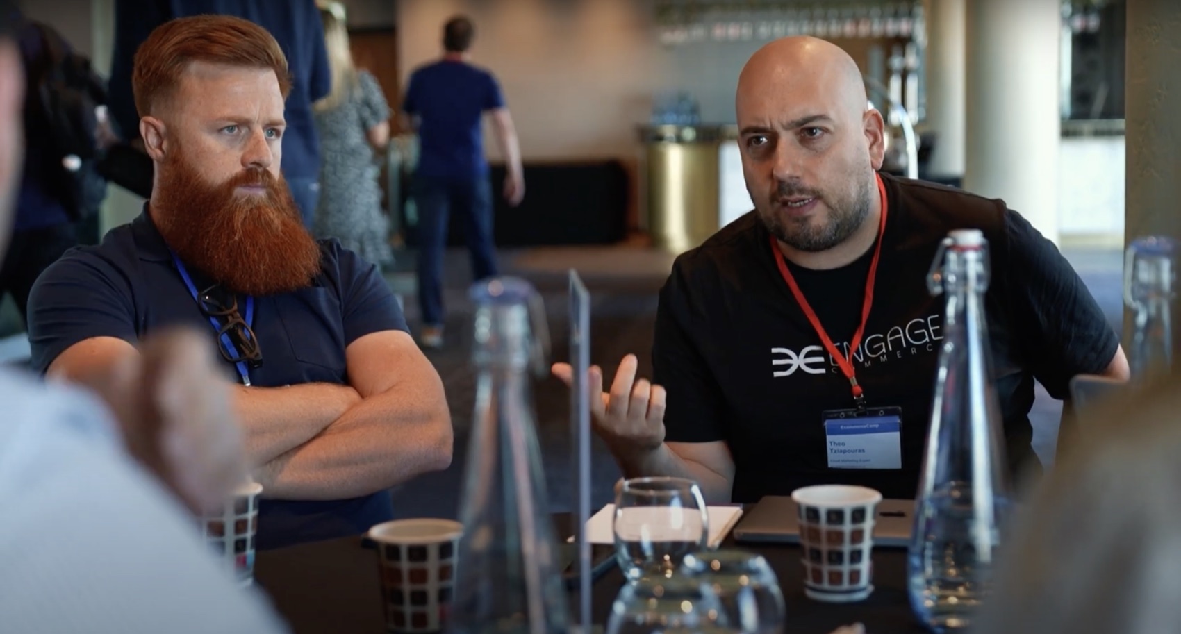 Theo Tziapouras in a black Engage Commerce t-shirt and red lanyard talking across a table to a bearded delegate in a navy polo shirt, water bottles and cups between them