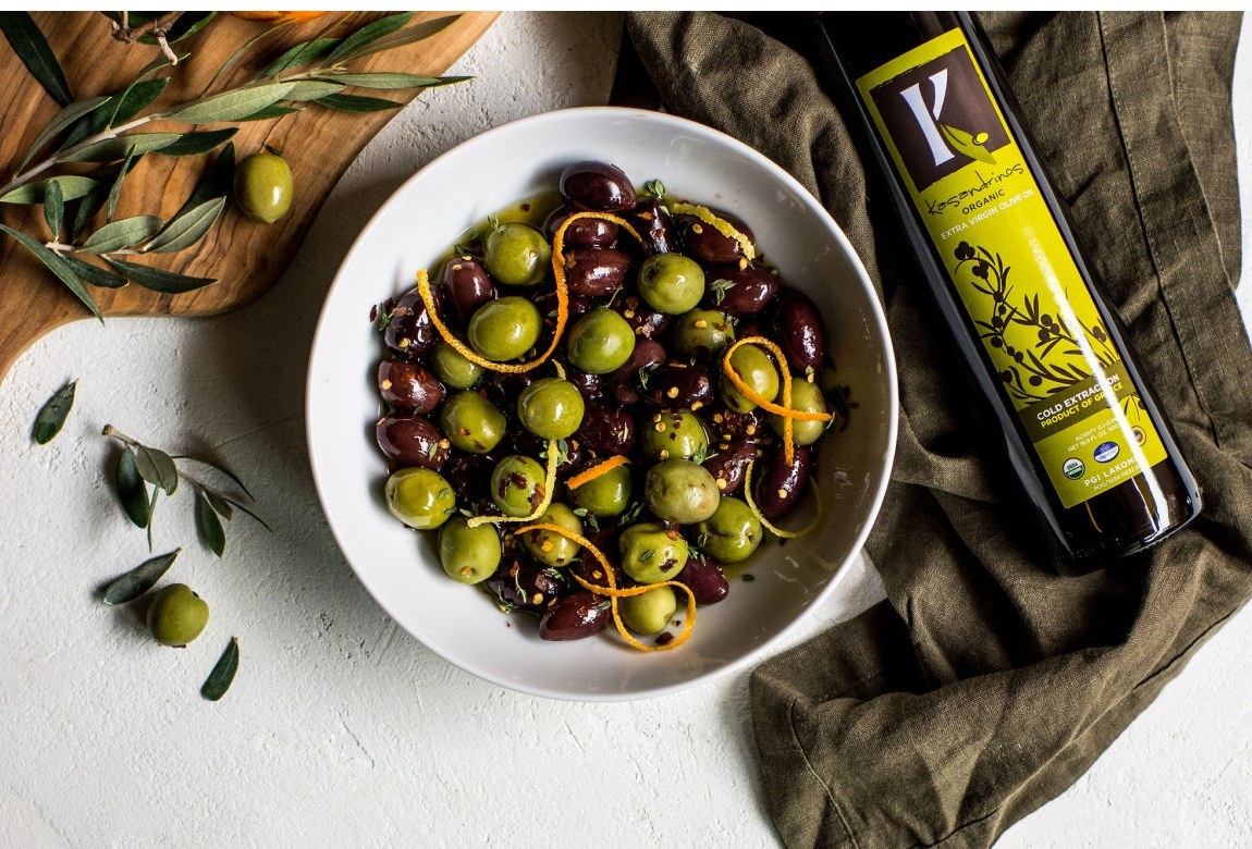 A bowl of marinated olives beside a bottle of Kasandrinos olive oil on a linen cloth