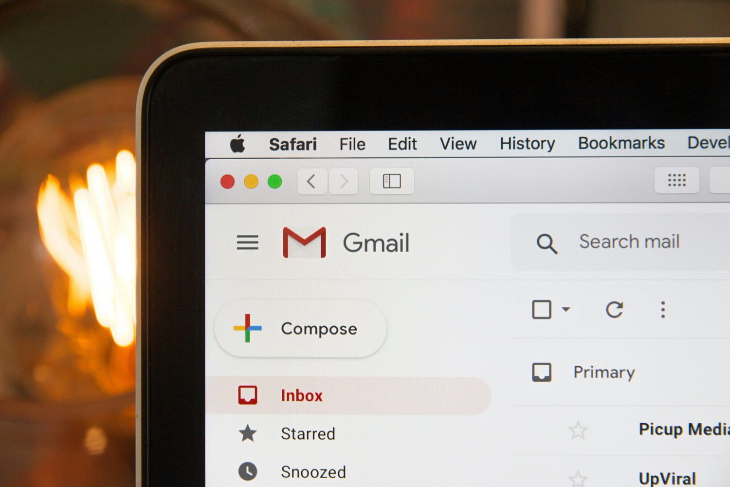 The corner of a laptop screen showing a Gmail inbox open in Safari, the Compose button and two sender names visible, warm lights blurred behind
