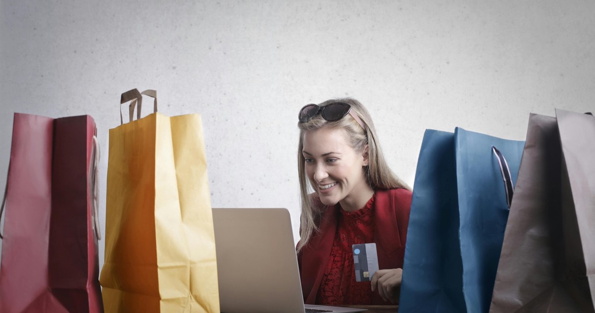 A smiling woman in red holds a bank card at a laptop, framed by colourful paper gift bags on either side