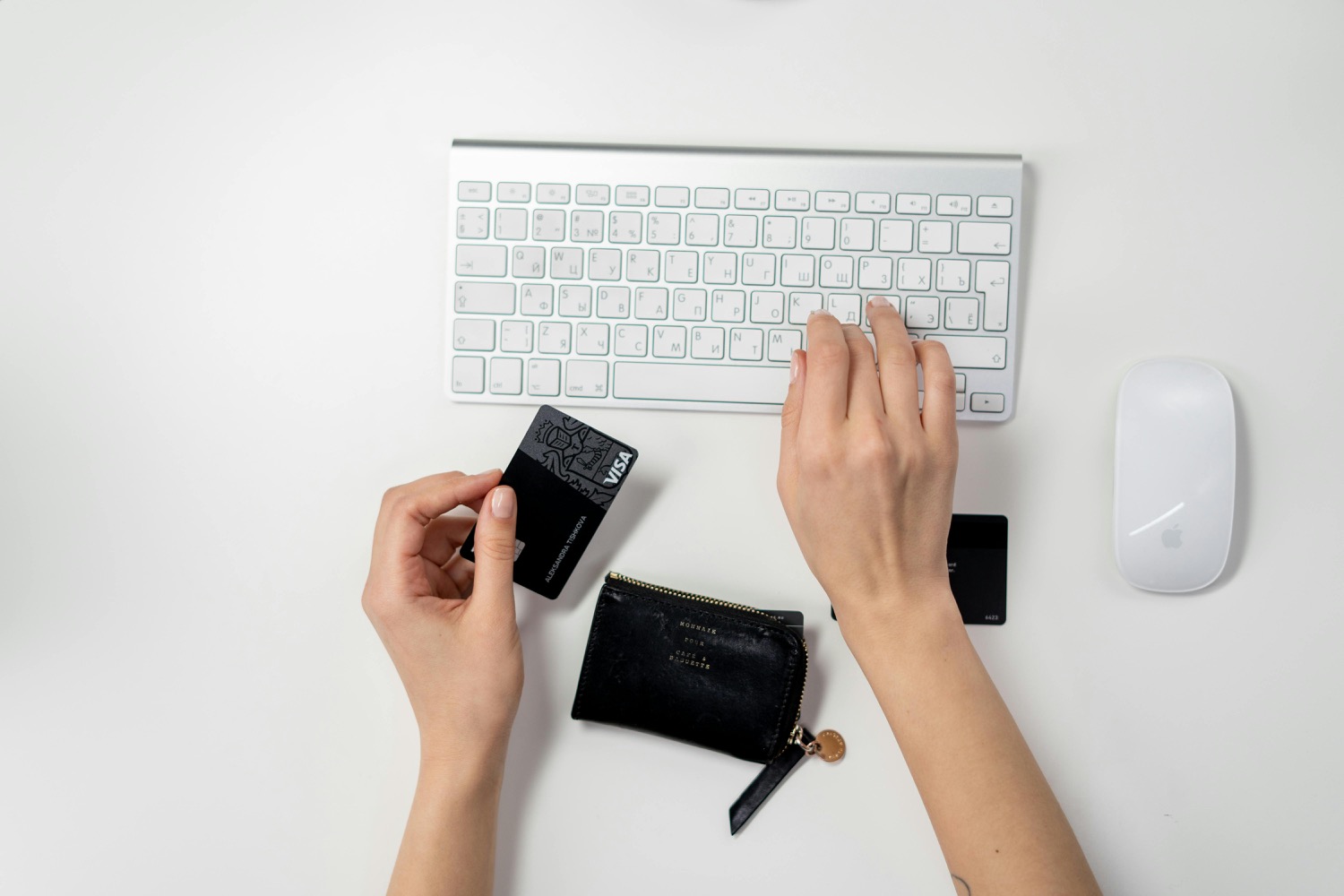 Overhead view of a woman's hands at a white keyboard, one holding a black bank card above a small zipped purse on the desk