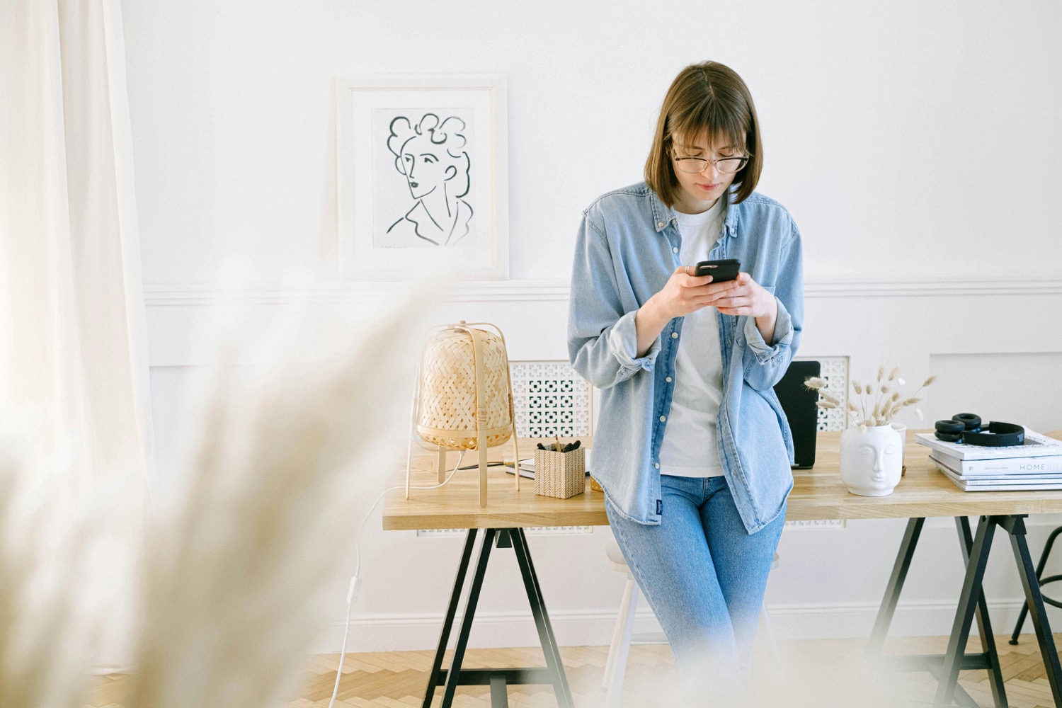 A woman in glasses and a denim shirt perches on a wooden trestle desk in a bright white home office, reading her phone