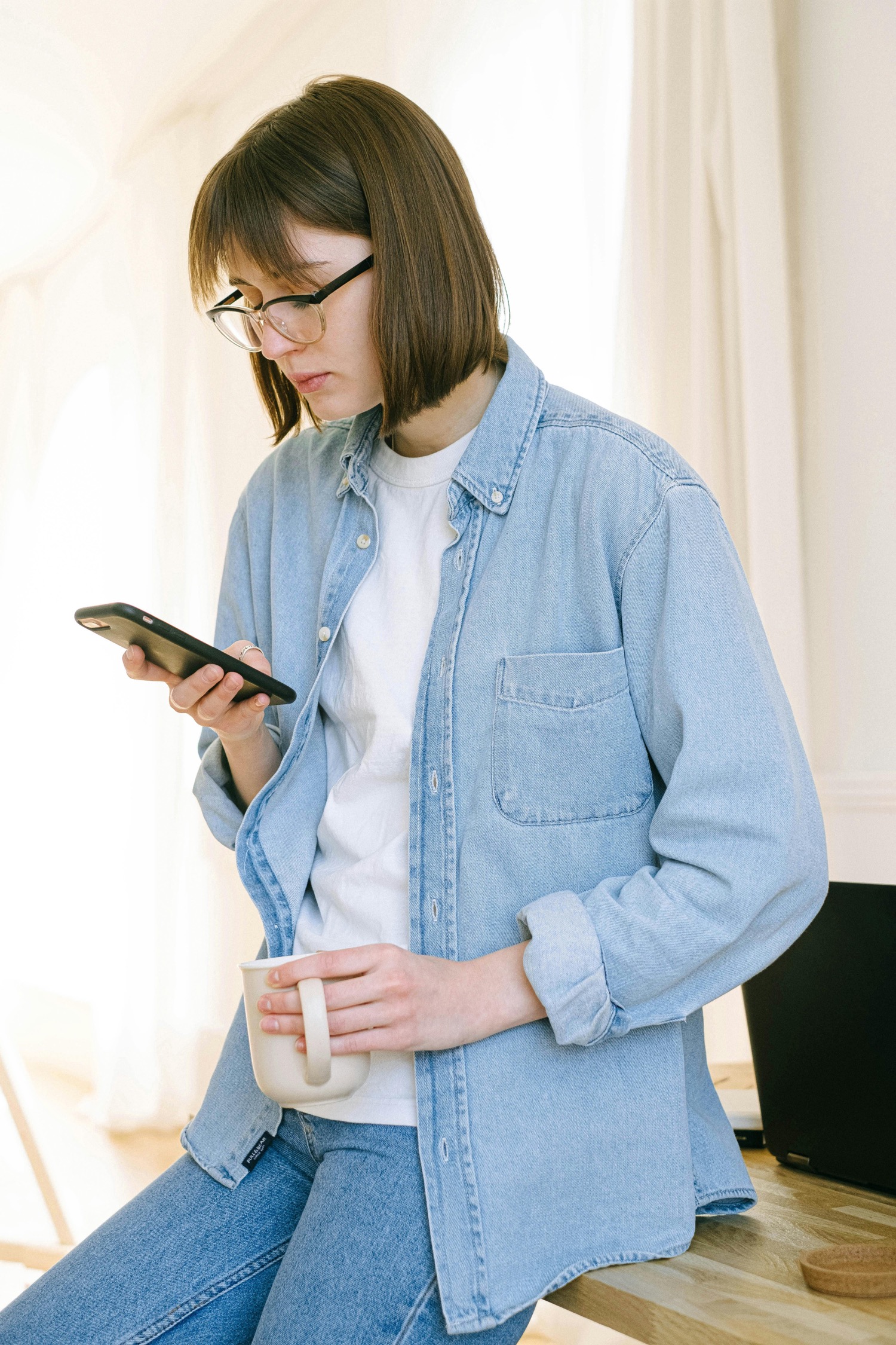 A woman in glasses and a light denim shirt holding a cream mug while reading something on her phone