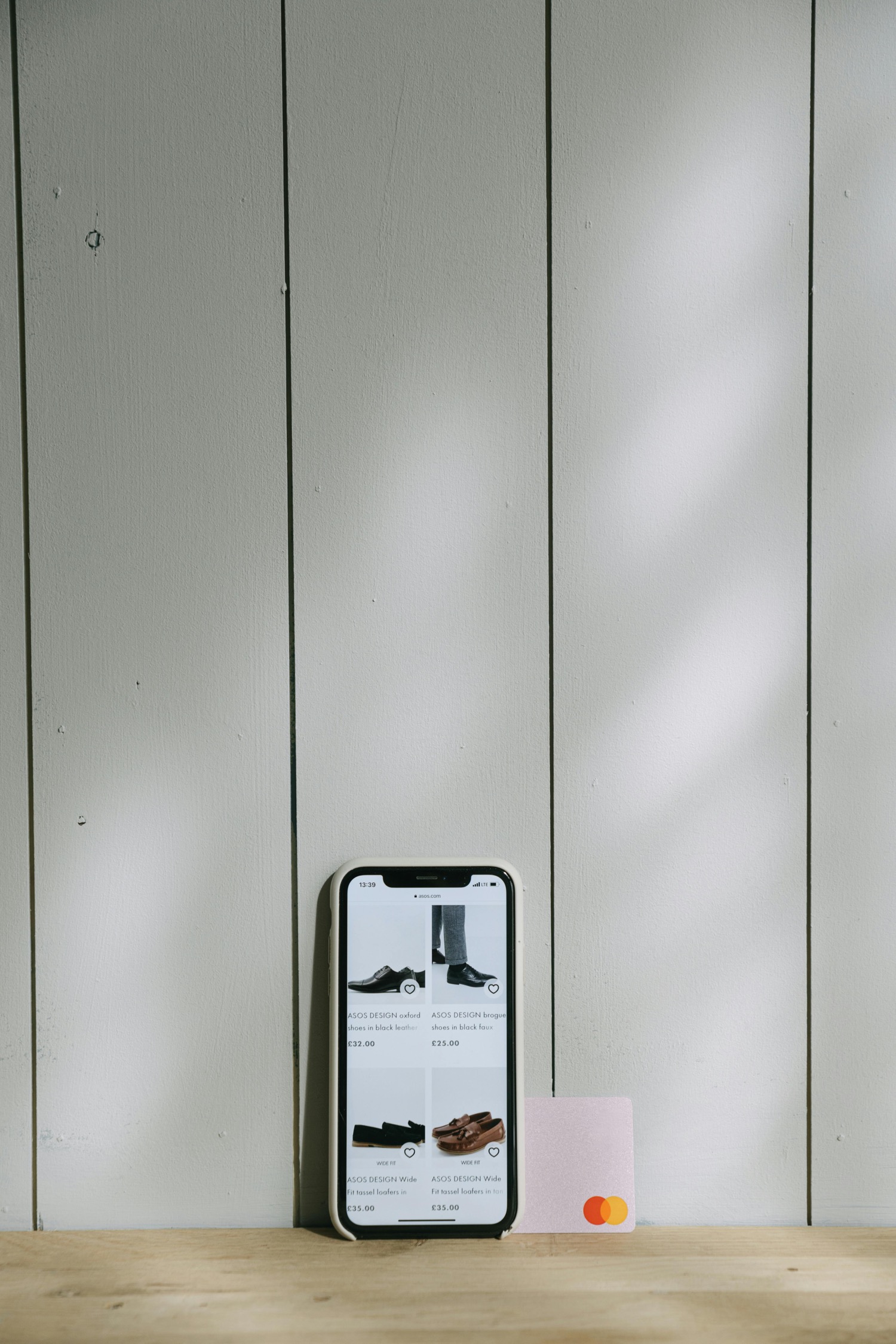 A phone propped against a painted wooden wall showing a shop listing page of black and tan shoes, a pink contactless card leaning beside it