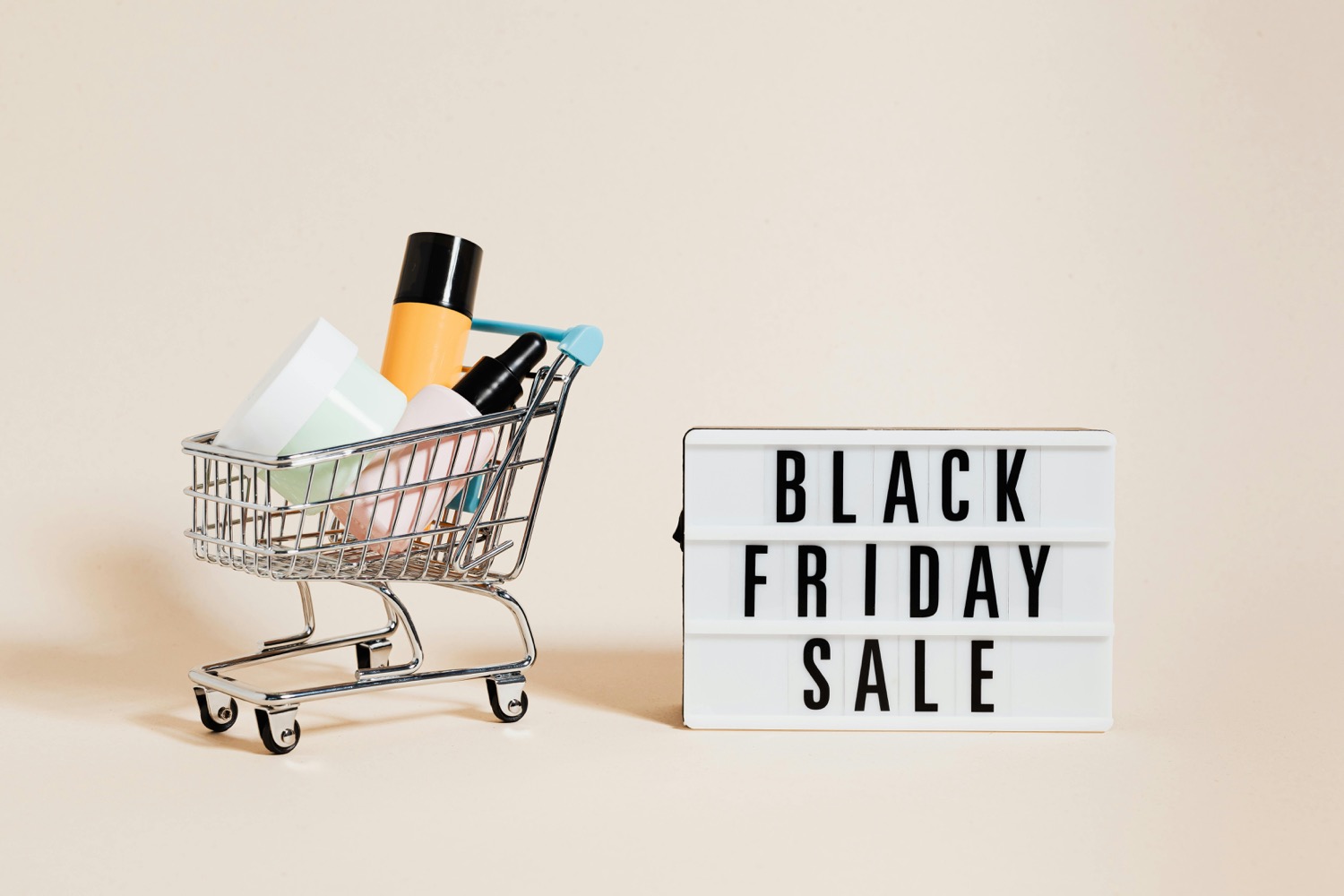 A miniature shopping trolley filled with cosmetics bottles beside a letterboard lightbox spelling out a Black Friday sale on a cream background