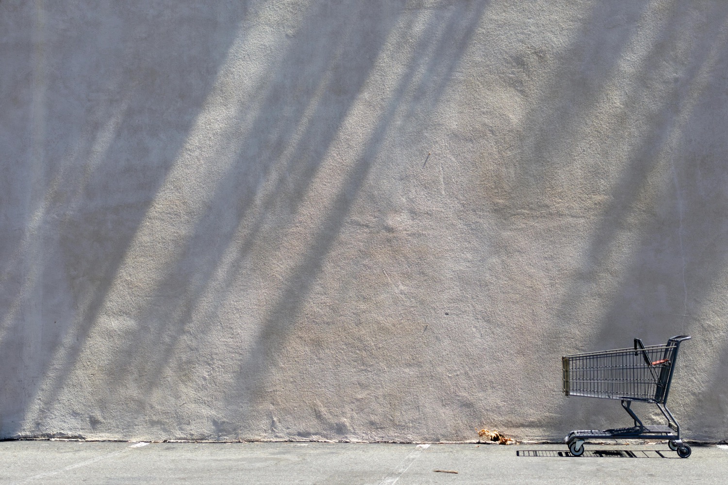 An empty metal shopping trolley standing alone against a vast sunlit concrete wall in an empty car park