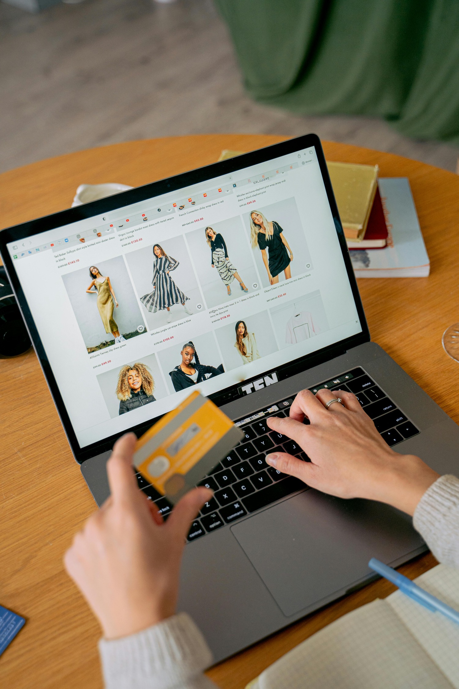 A shopper at a laptop with a card in her hand, an ecommerce category page of dresses open on screen with prices under every tile