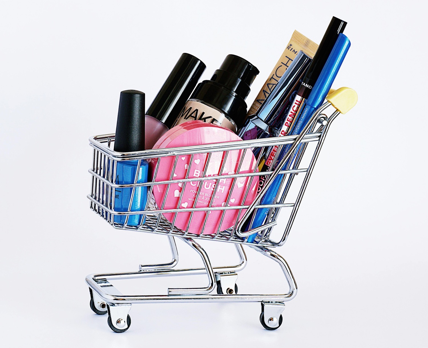 A miniature chrome shopping trolley filled with cosmetics, including a pink blusher compact, a foundation bottle and eyeliner pencils, against a plain white background
